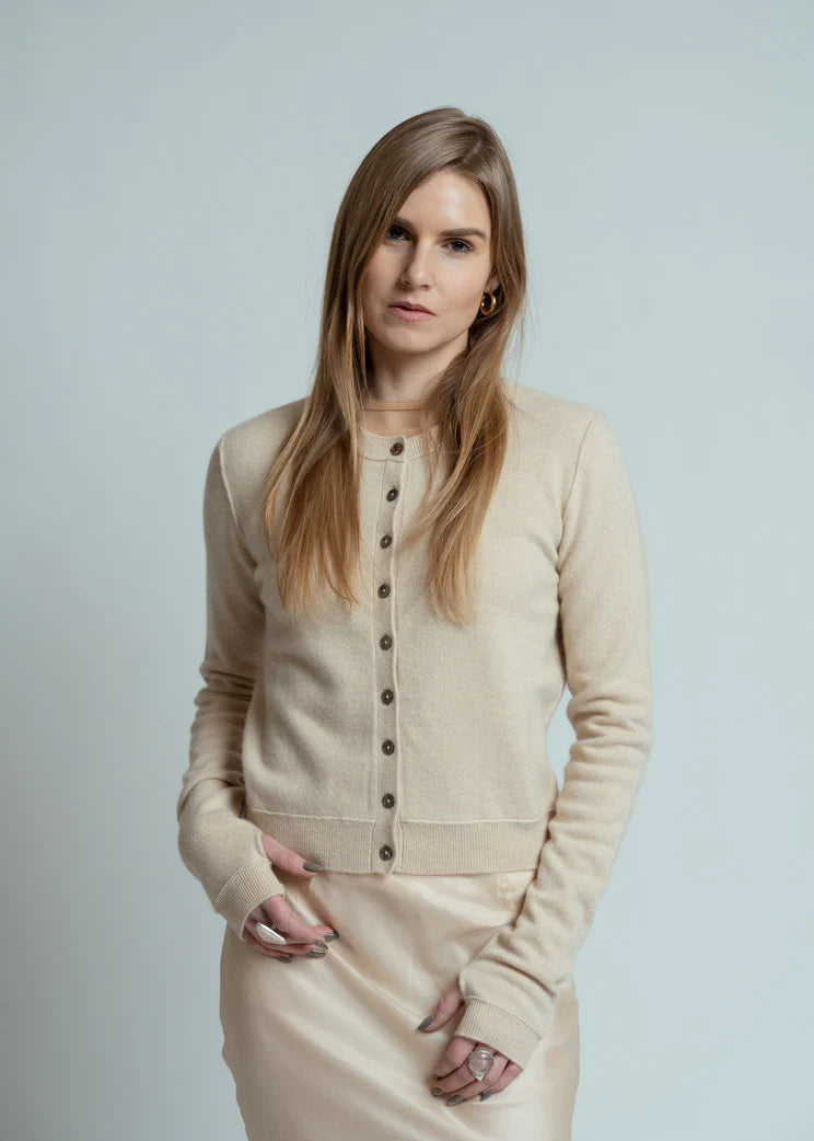 A woman with long blonde hair and a neutral expression stands against a plain light background, wearing the Brazeau Tricot Royal All Thumbs Cardigan in beige with a button front, paired with a matching skirt.