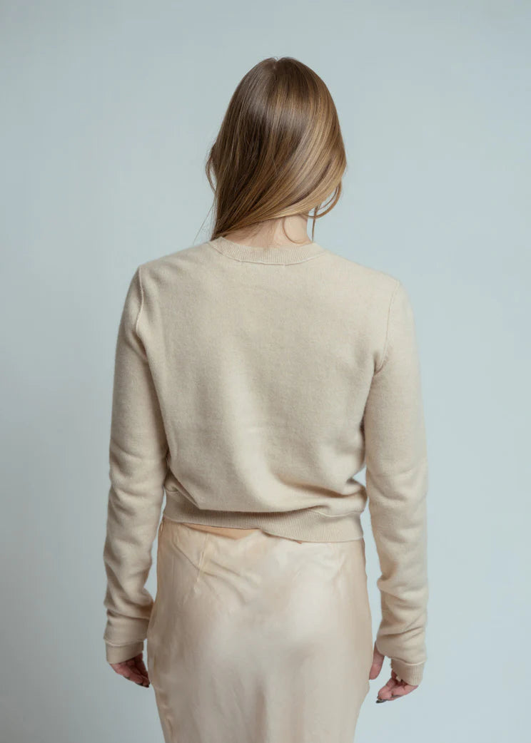 A person with long, straight light brown hair stands with their back to the camera, wearing the Brazeau Tricot Royal All Thumbs Cardigan with button front and a cream skirt against a plain light background.