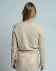 A person with long, straight light brown hair stands with their back to the camera, wearing the Brazeau Tricot Royal All Thumbs Cardigan with button front and a cream skirt against a plain light background.
