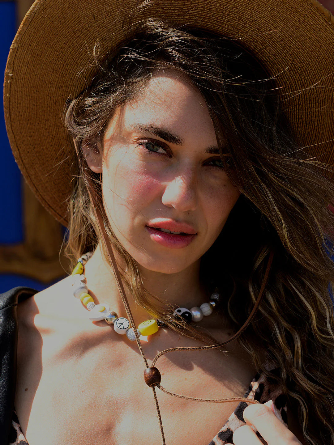 A woman with wavy hair and a wide-brimmed hat wears the Venessa Arizaga Sunny Side Necklace 16" with pavé rhinestone charms, gazing at the camera in sunlight, her patterned top and hair partly covering one eye.