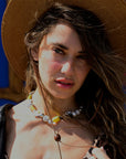 A woman with wavy hair and a wide-brimmed hat wears the Venessa Arizaga Sunny Side Necklace 16" with pavé rhinestone charms, gazing at the camera in sunlight, her patterned top and hair partly covering one eye.