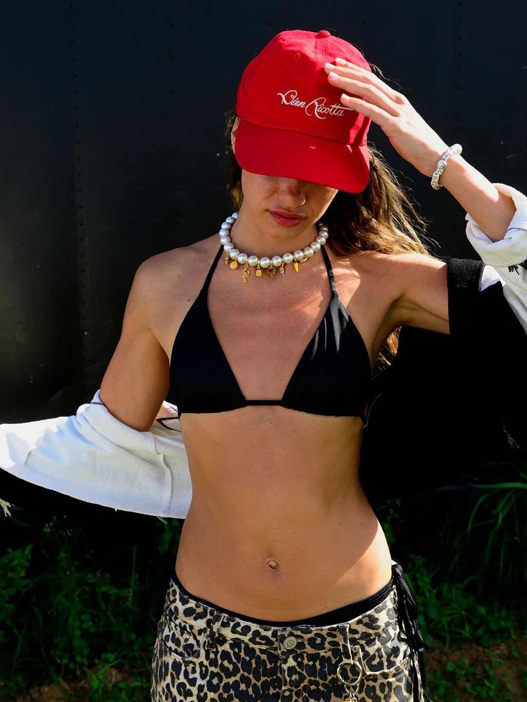 A person outdoors with greenery behind them wears a red cap, black bikini top, leopard print bottoms, and a Venessa Arizaga Brunch by the Beach Necklace 16", with a jacket draped over their arms and head slightly bowed.