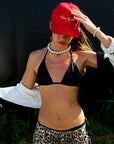 A person outdoors with greenery behind them wears a red cap, black bikini top, leopard print bottoms, and a Venessa Arizaga Brunch by the Beach Necklace 16", with a jacket draped over their arms and head slightly bowed.
