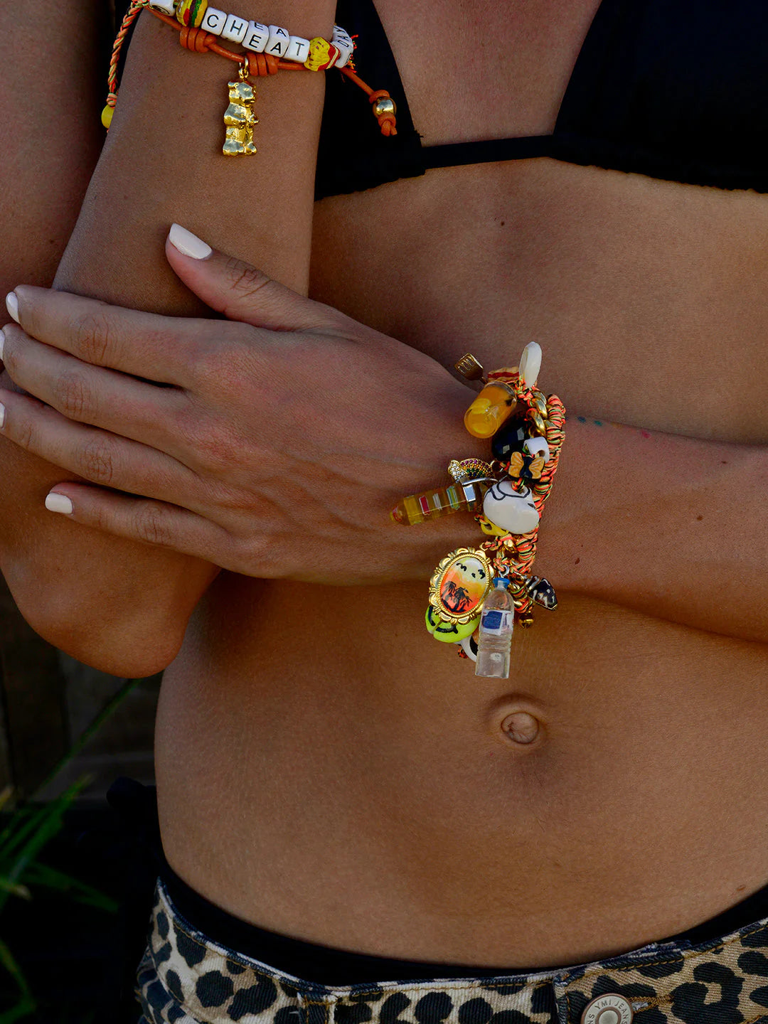 A person with light brown skin, in a black bikini top and leopard print bottoms, crosses their arms adorned with Venessa Arizaga’s Tropical Treats Gold Plated bracelet featuring colorful charms and "CHEAT" letter beads. Their nails are painted white.