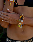A person with light brown skin, in a black bikini top and leopard print bottoms, crosses their arms adorned with Venessa Arizaga’s Tropical Treats Gold Plated bracelet featuring colorful charms and "CHEAT" letter beads. Their nails are painted white.