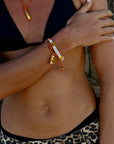 A person in a black bikini top and leopard print bottoms stands with one arm crossed, wearing the Venessa Arizaga Bracelet 6"—an adjustable charm bracelet with white beads spelling "CHEERS" and small gold charms—plus a matching necklace.
