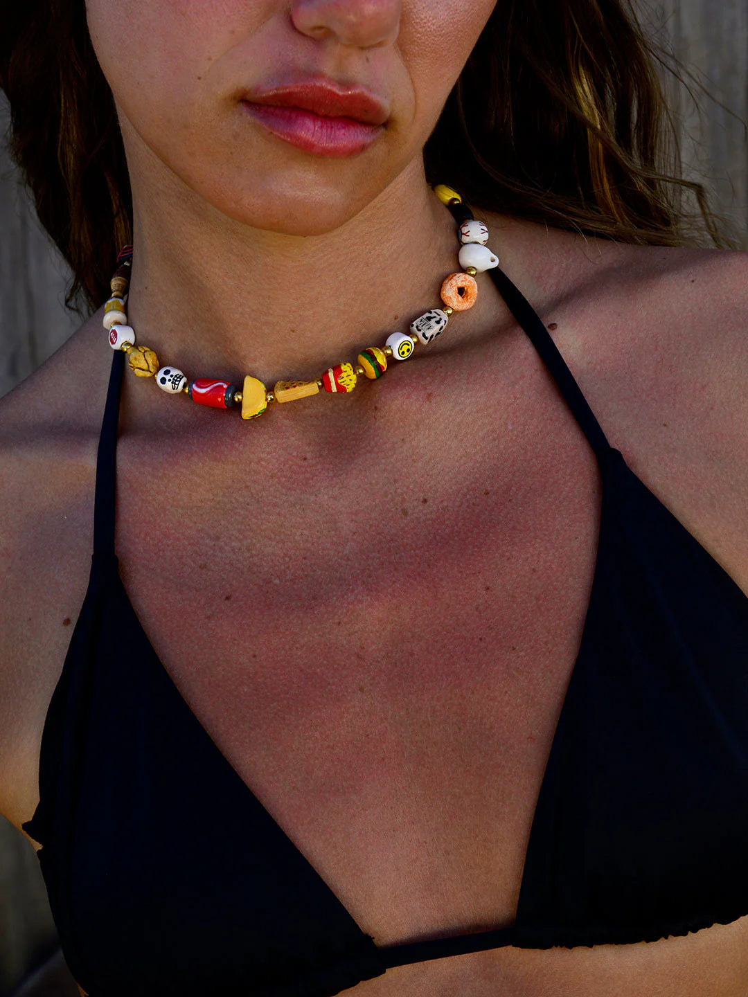 A woman wears a black bikini top and the Venessa Arizaga Breakfast Lunch Dinner Necklace 14", featuring playful food charms. Tan lines and slight redness are visible on her chest and shoulders.