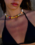 A woman wears a black bikini top and the Venessa Arizaga Breakfast Lunch Dinner Necklace 14", featuring playful food charms. Tan lines and slight redness are visible on her chest and shoulders.