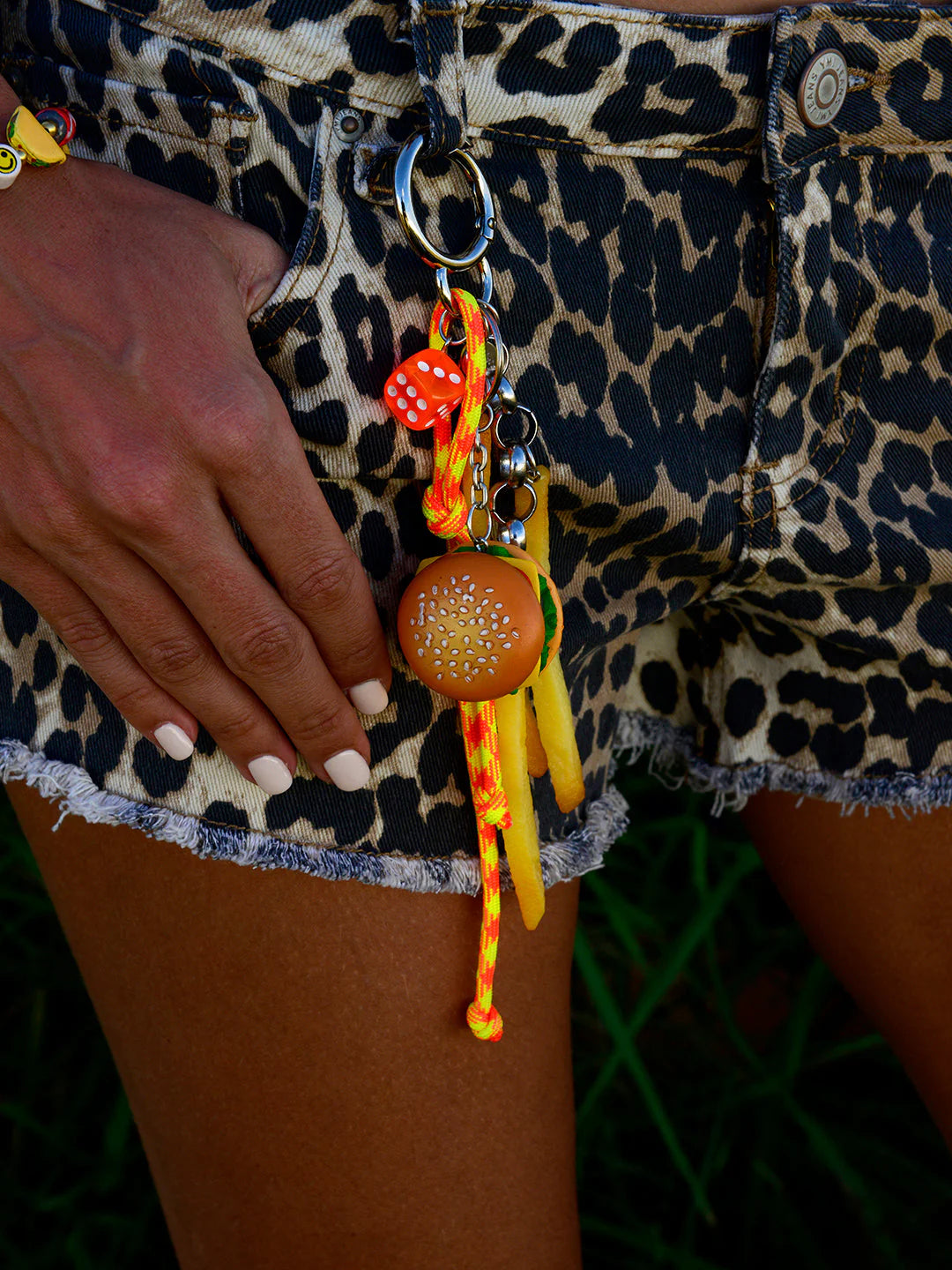 A person in leopard print denim shorts holds the Venessa Arizaga Bag Key Leash, decorated with food charms—a hamburger, yellow fries, an orange dice, and colorful trinkets. Their manicured nails add to the playful style.