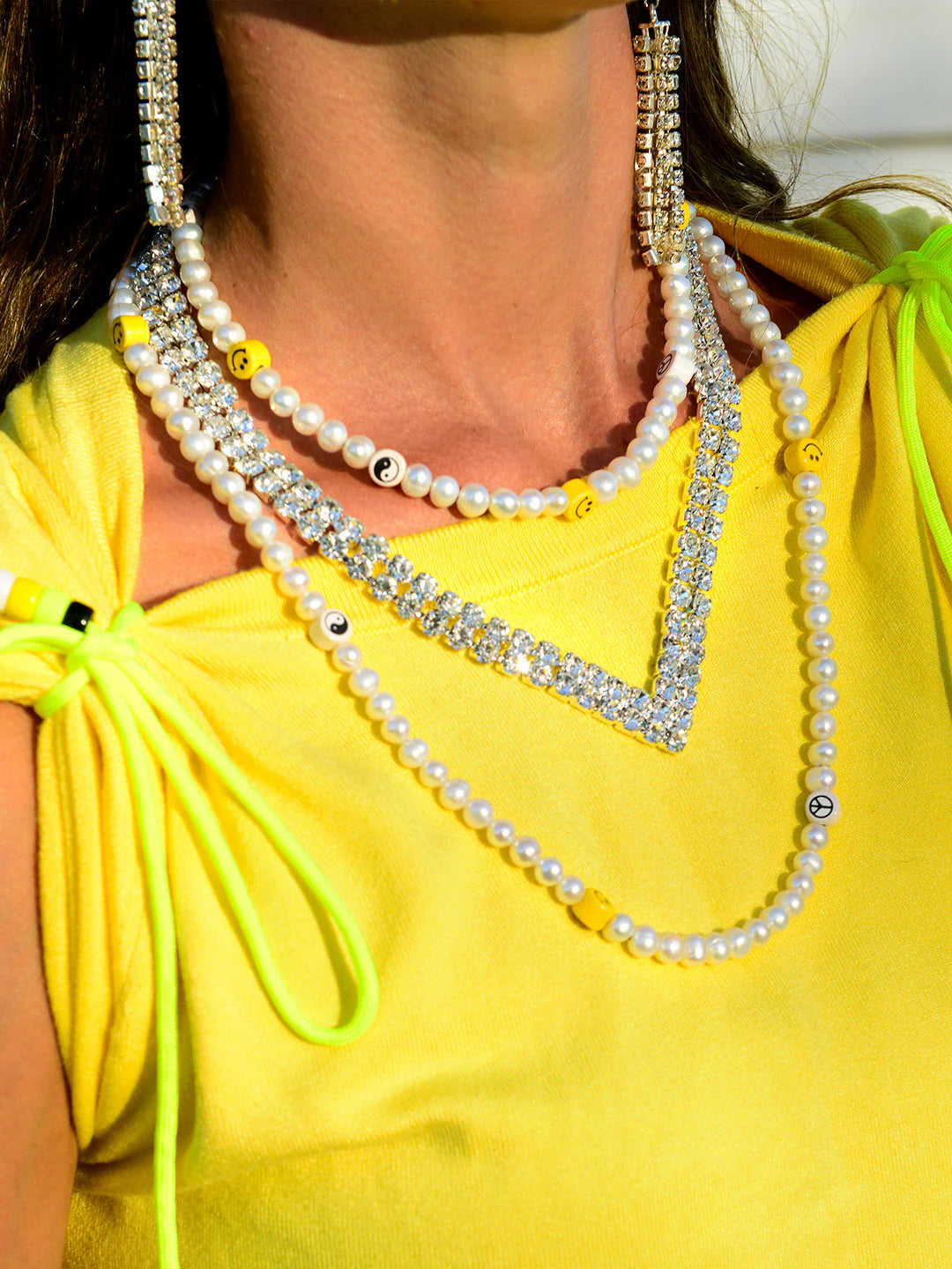 A person wears layered freshwater pearl and rhinestone necklaces, including Venessa Arizaga's Happy Dream Necklace 16-26", featuring yellow smiley face and peace sign beads, over a bright yellow sleeveless top with gathered shoulder ties.