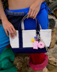 A person in colorful pants holds a Venessa Arizaga Bag Key Leash—a white and navy tote adorned with playful food charms, like a pink chocolate bar, donut bag charm, and fuzzy ball—standing by a stone wall.