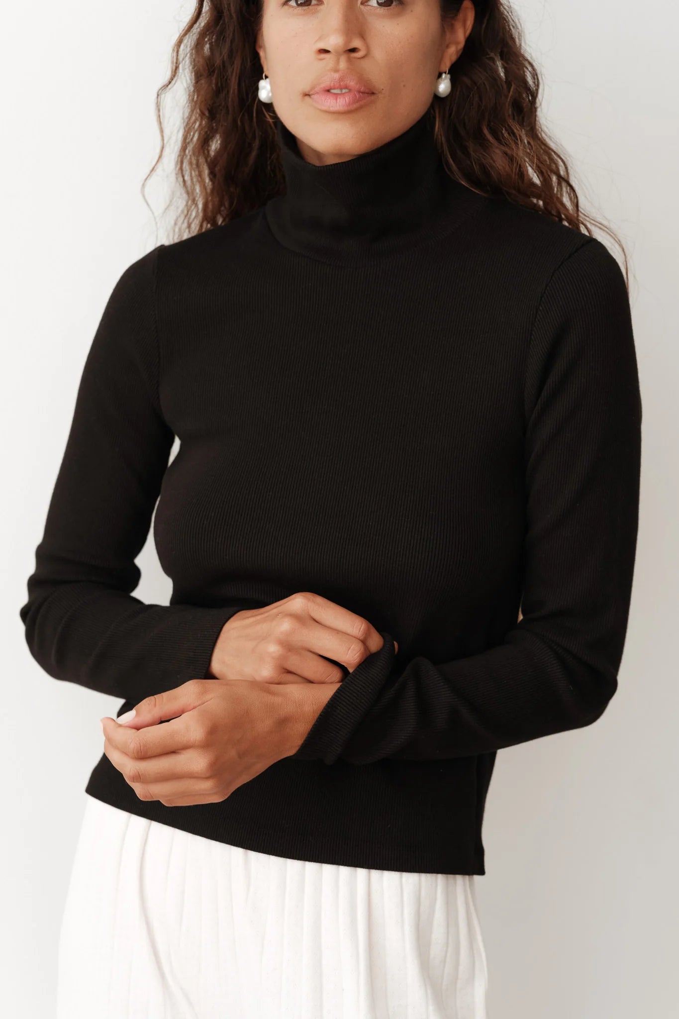 A person wearing "The Rib Turtleneck" by Donni in black, paired with white pleated pants, stands against a plain background. They have long hair and are adjusting the sleeve of their top, showcasing the fine rib texture.