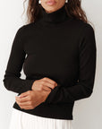 A person wearing "The Rib Turtleneck" by Donni in black, paired with white pleated pants, stands against a plain background. They have long hair and are adjusting the sleeve of their top, showcasing the fine rib texture.