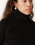 A person with long curly hair is wearing "The Rib Turtleneck" by Donni and pearl earrings, set against a plain, light-colored background.