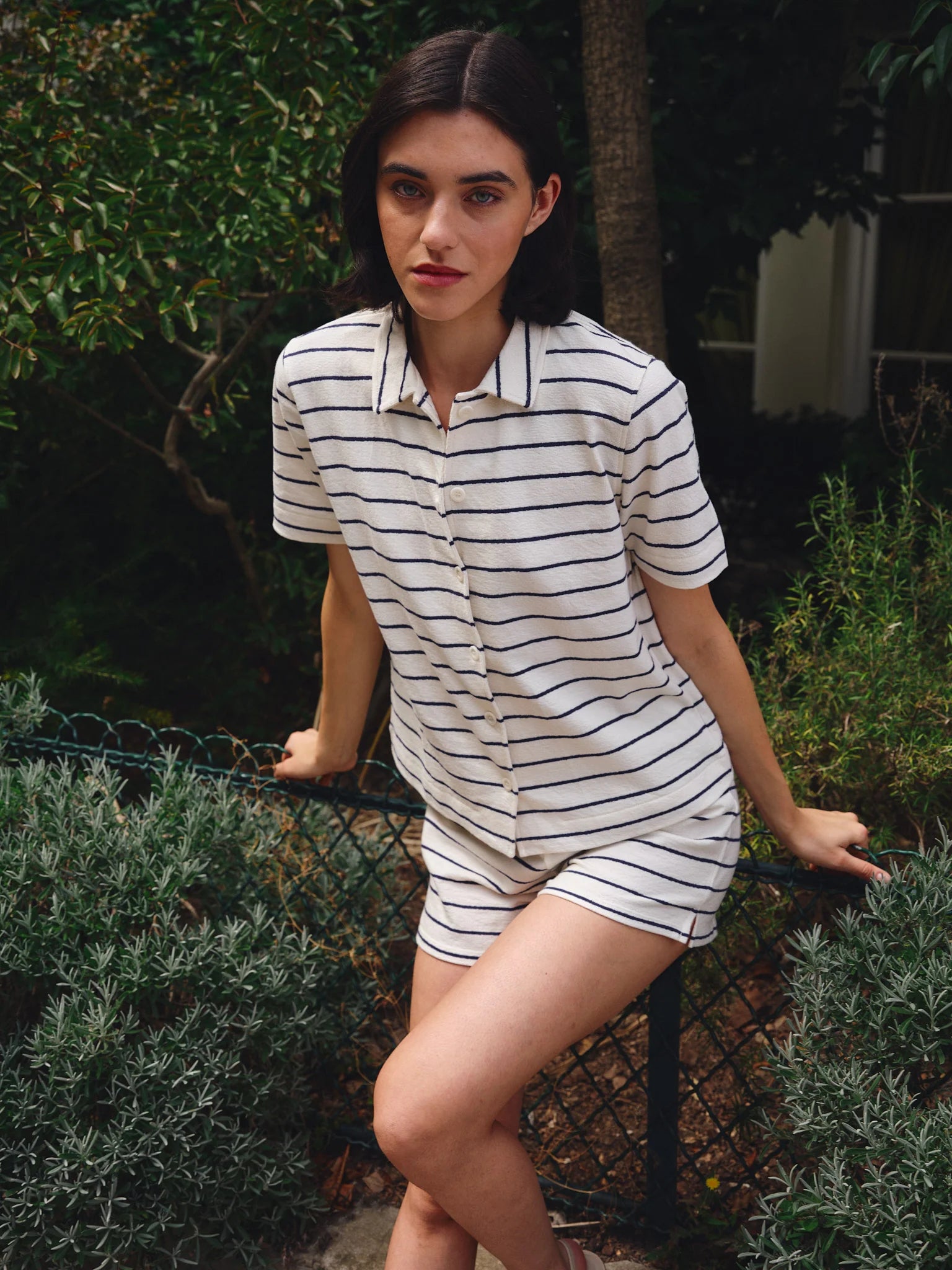 A woman with dark hair, wearing a short-sleeved white button-up shirt and Ines De LA Fressange Lucas Shorts, sits on a fence in a garden surrounded by lush green plants and trees.