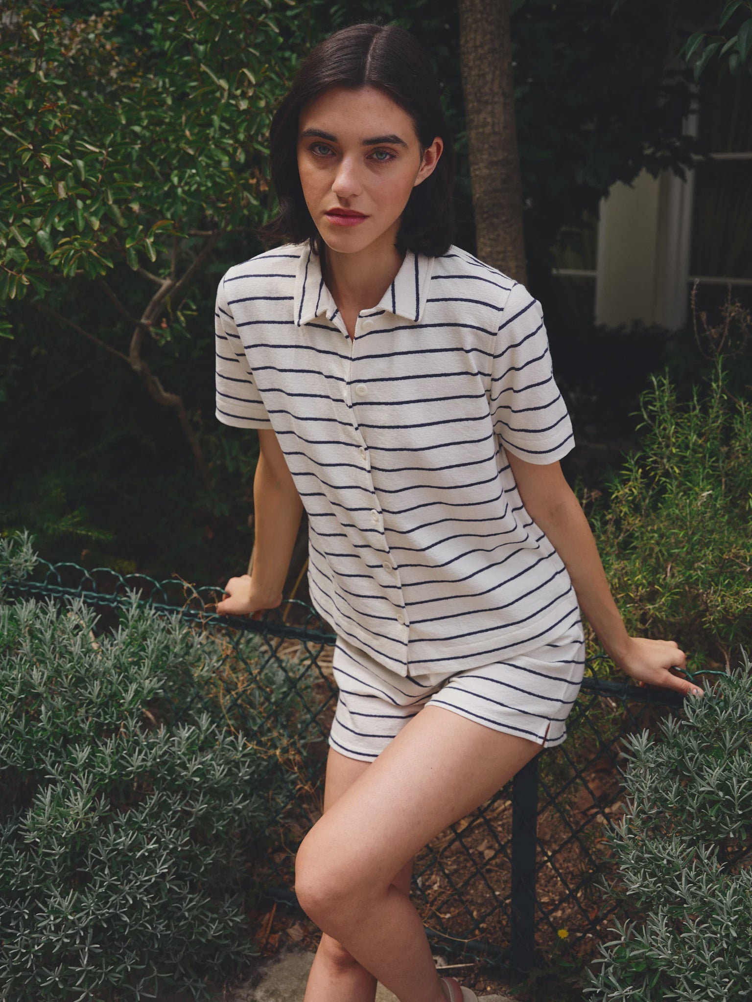 A dark-haired woman stands outdoors by a fence amid green foliage, wearing the Ines De LA Fressange Nicia Shirt and matching shorts.