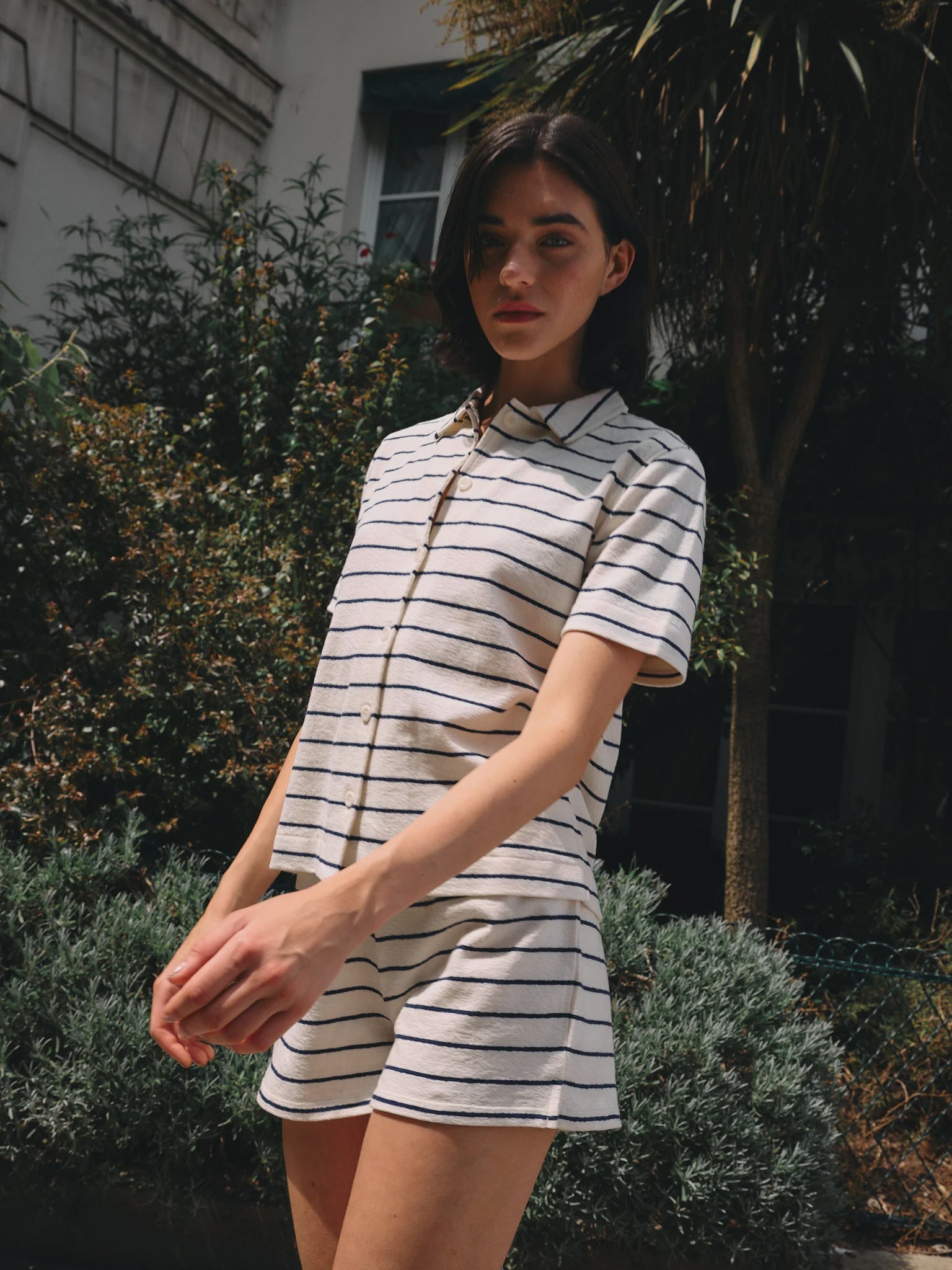 A person with short dark hair wears the Ines De LA Fressange Lucas Short, a light-colored, short-sleeved, striped romper made from organic cotton, while standing outdoors in front of greenery and a building.