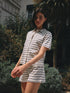 Standing outdoors before lush greenery, a person with short dark hair wears the Ines de la Fressange Lucas Short, a collared, short-sleeved striped romper crafted from organic cotton, gazing slightly to the side in natural sunlight.