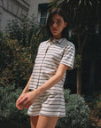 A person with short dark hair wears the Ines De LA Fressange Lucas Short, a light-colored, short-sleeved, striped romper made from organic cotton, while standing outdoors in front of greenery and a building.