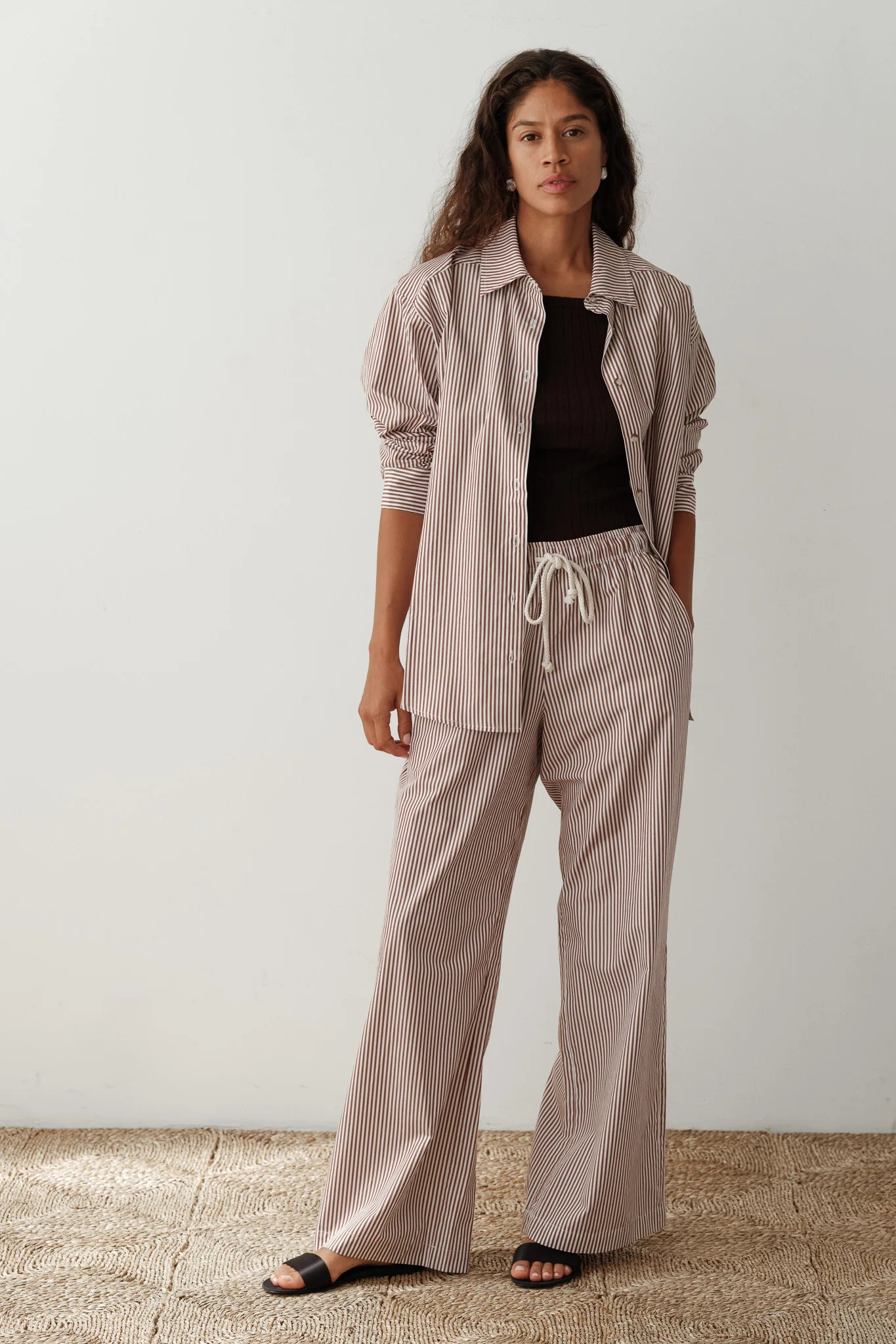 Against a plain backdrop, a person with long, wavy hair poses with one hand in their pocket, wearing Donni's brown and white striped cotton poplin shirt and The Stripe Pop Pant. The wide-leg fit is enhanced by a black top and sandals.