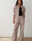 Against a plain backdrop, a person with long, wavy hair poses with one hand in their pocket, wearing Donni's brown and white striped cotton poplin shirt and The Stripe Pop Pant. The wide-leg fit is enhanced by a black top and sandals.