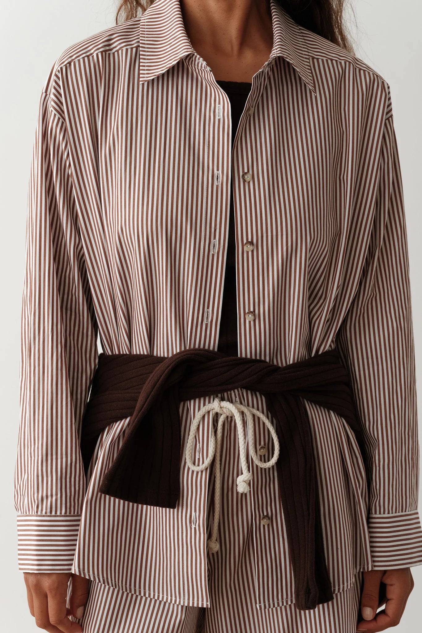 A person wears The Stripe Pop Shirt by Donni, characterized by its brown and white striped cotton poplin design, paired with matching pants. A dark brown sweater is tied around their waist, and the pants are complemented by a white drawstring. This stylish ensemble, crafted in LA, stands out against the plain, light-colored background.