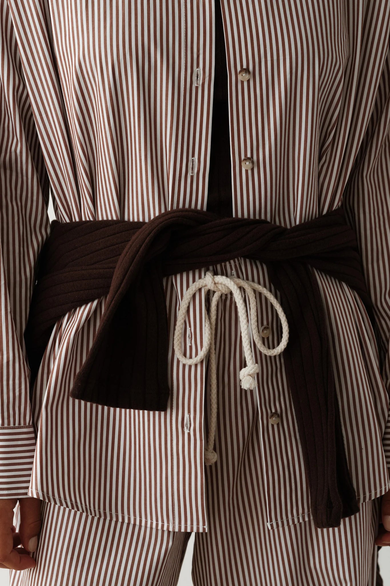 A person is wearing The Stripe Pop Shirt by Donni, which showcases vertical brown and white stripes crafted from cotton poplin. The shirt features tortoise-like buttons and is styled with pants, along with a dark sweater tied around the waist using a white rope.