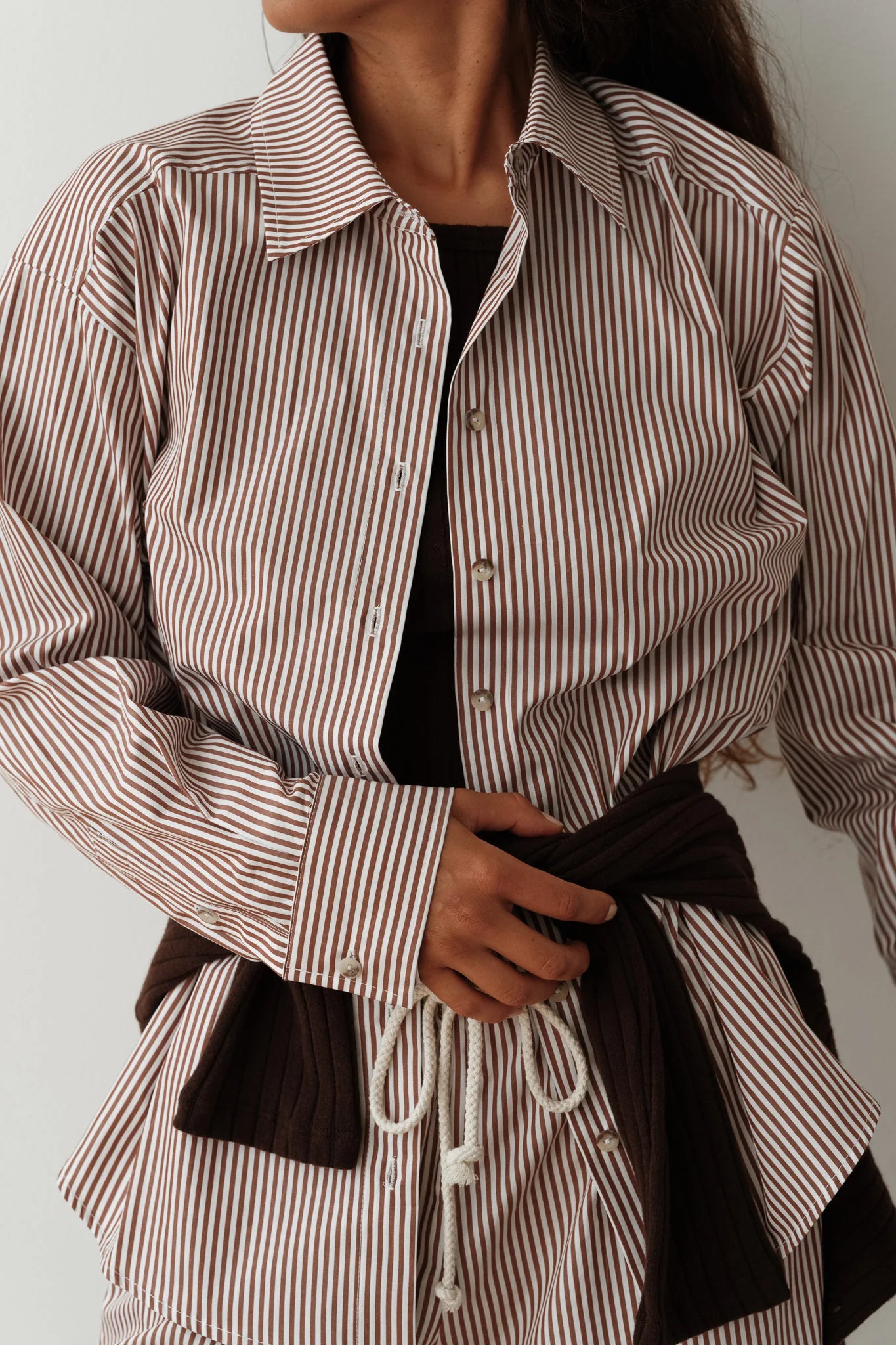 A person is dressed in The Stripe Pop Shirt by Donni, a brown and white cotton poplin button-up featuring tortoise-like buttons, layered over a dark top. They also have a brown sweater tied around their waist and are placing their hand on their abdomen, with their face not visible.