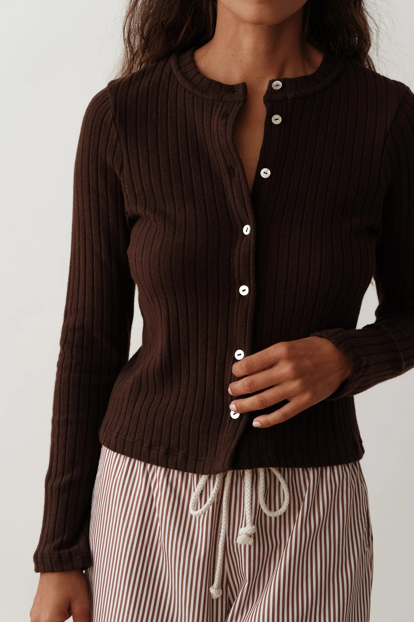 A woman wearing Donni's The Sweater Rib Card in brown, featuring white buttons, is adjusting her top. She complements it with striped drawstring pants against a plain white background.