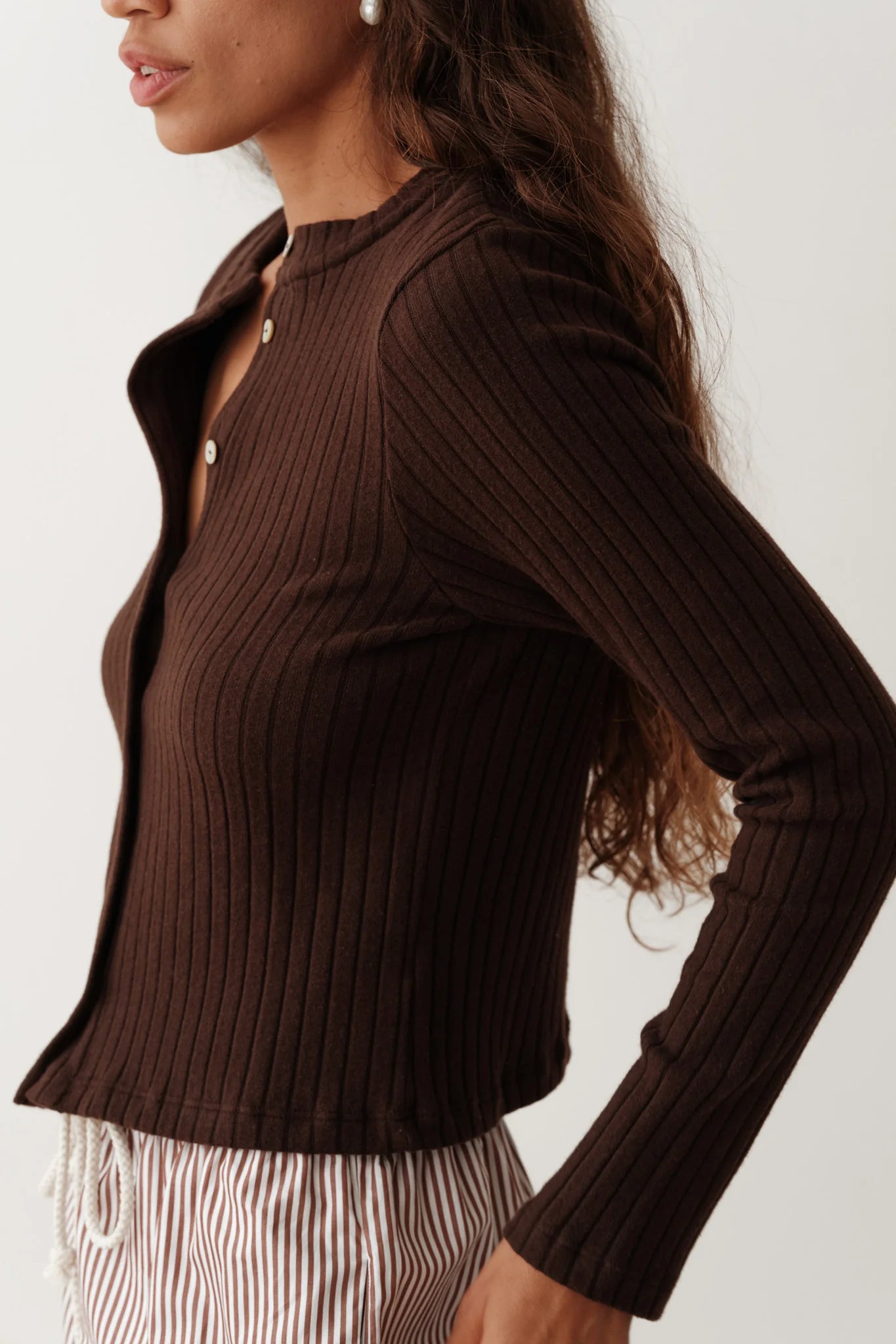 A person with long, wavy hair is wearing Donni's The Sweater Rib Card, leaving a few buttons undone for a relaxed look. They are also dressed in striped pants against a plain, neutral background.