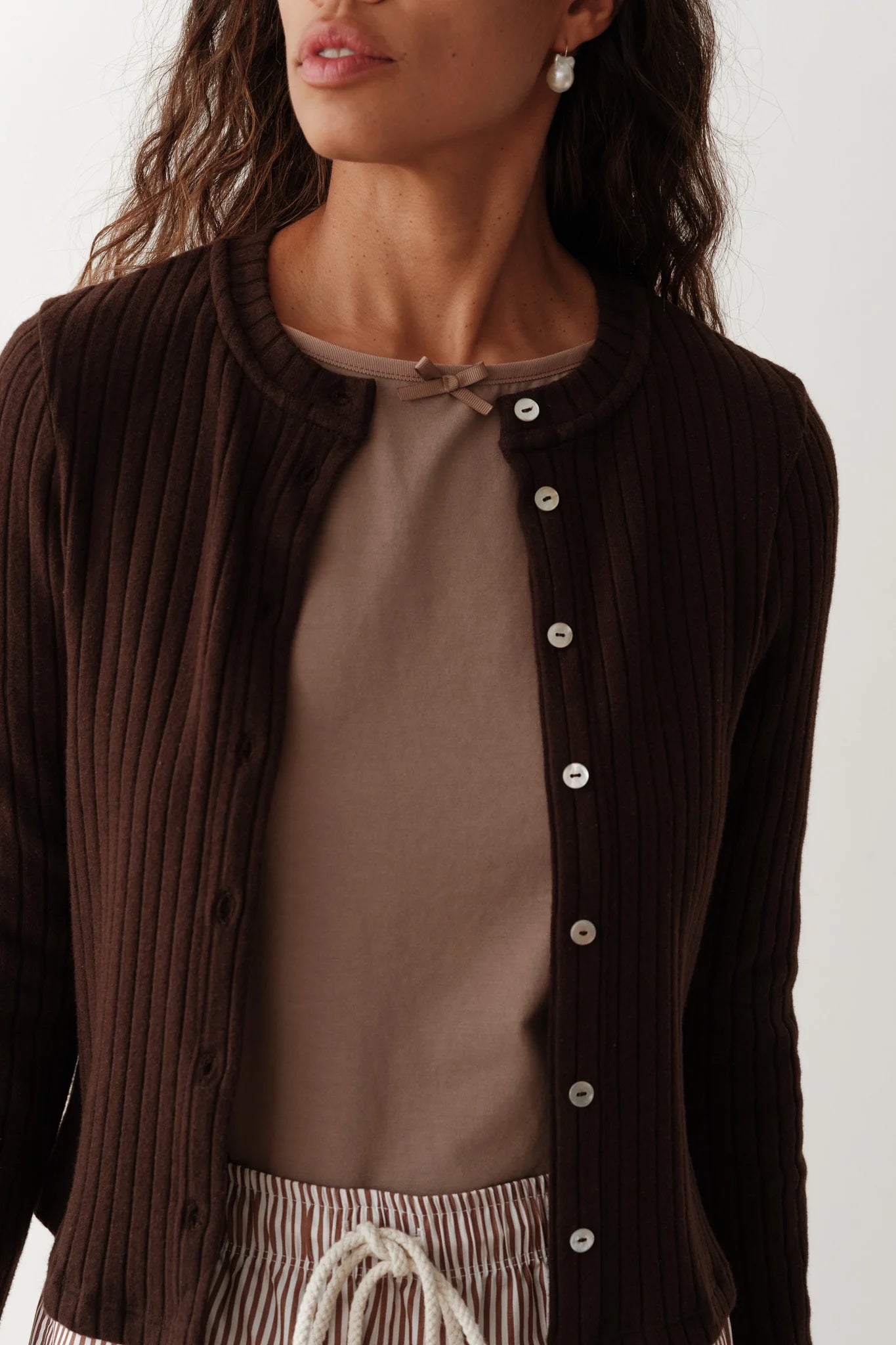 A woman is wearing "The Sweater Rib Card" by Donni, a cozy dark brown cardigan, over a taupe top with a small bow at the neckline. She has pearl earrings, and her wavy hair falls past her shoulders. The cardigan is unbuttoned, revealing striped pants with a drawstring.