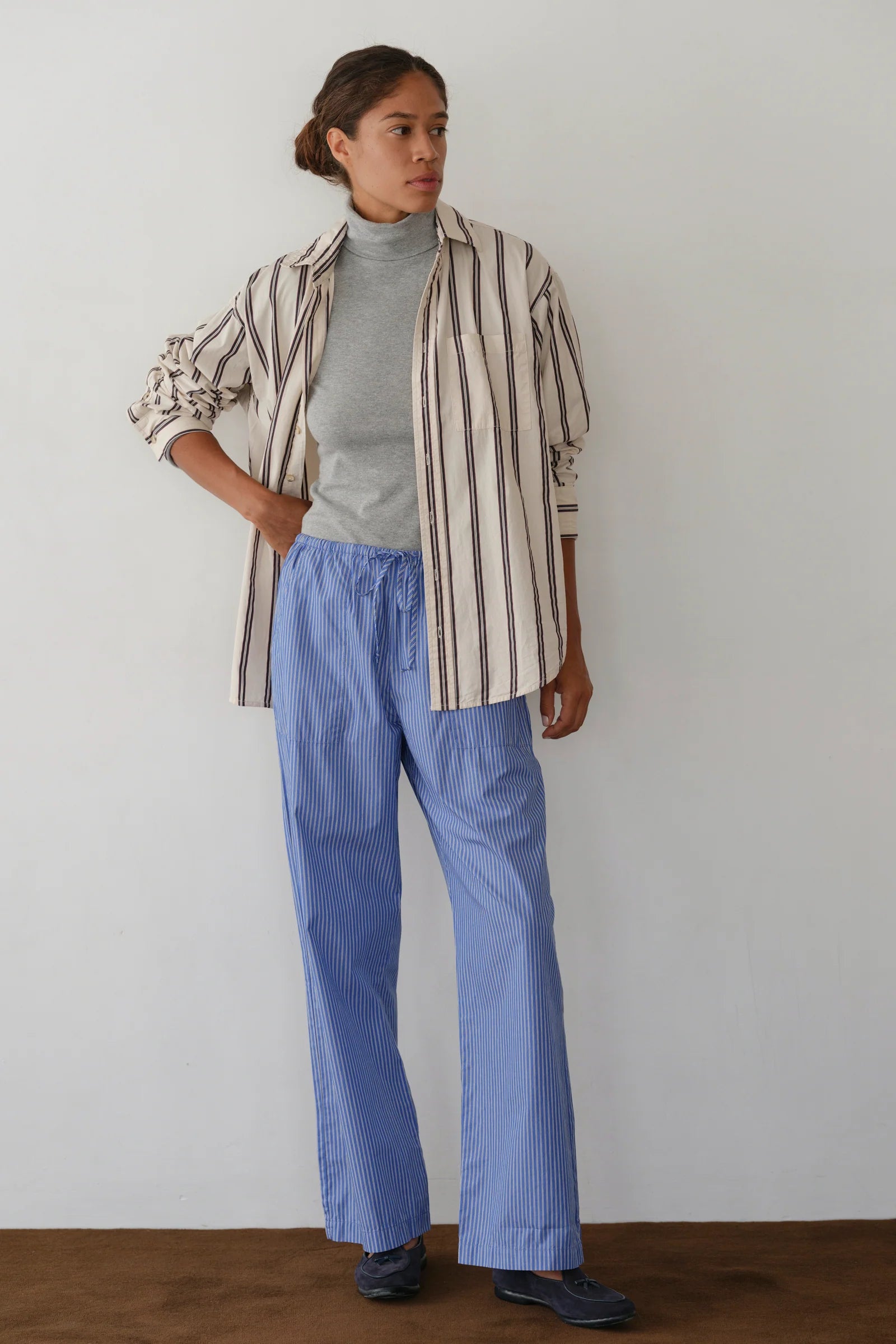 A person with short hair stands casually against a plain wall wearing Donni’s The Stripe Pop Drawstring Pant, a loose striped Cotton Poplin button-up shirt over a gray turtleneck, and dark shoes.