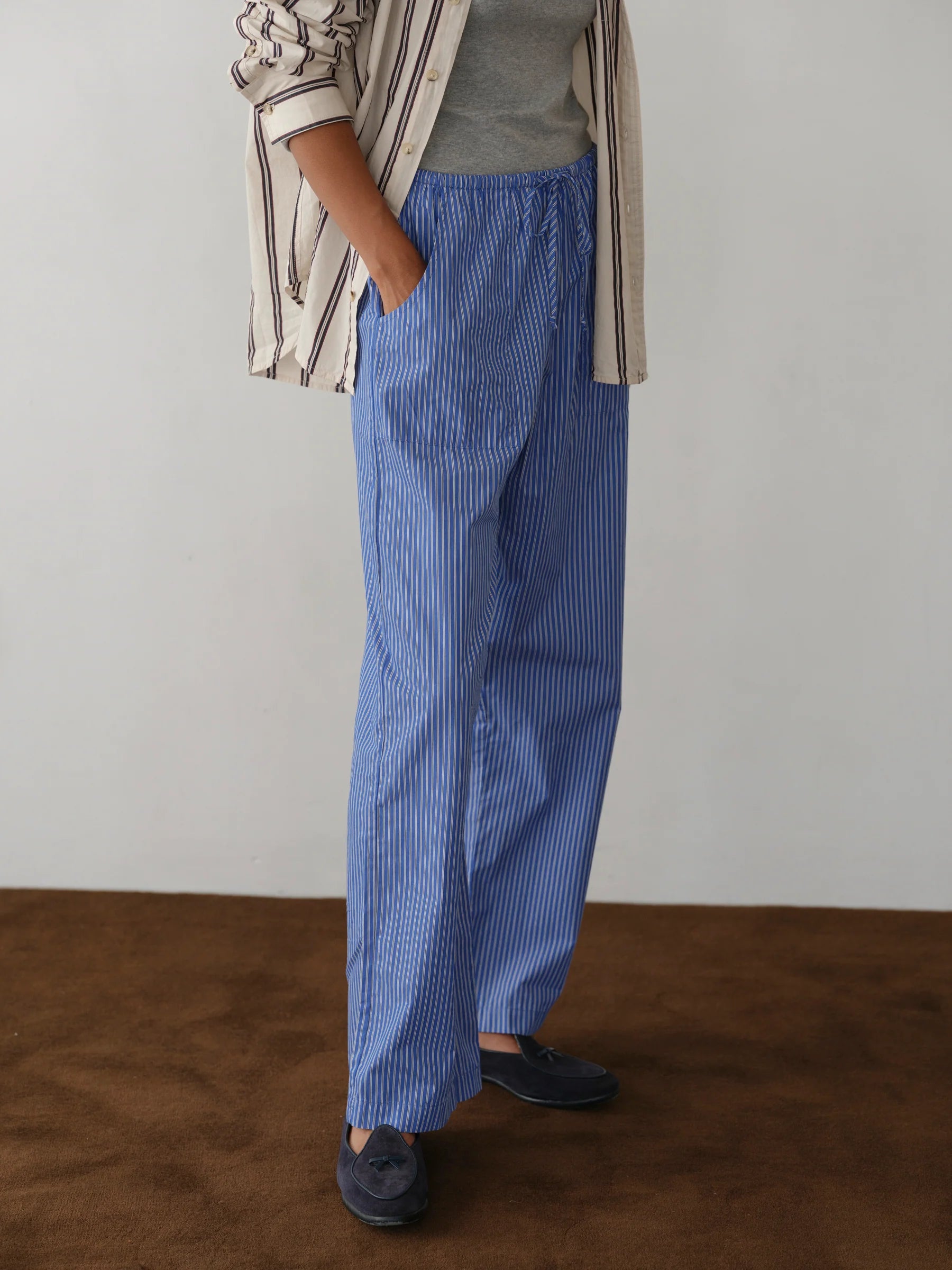 Wearing Donni’s The Stripe Pop Drawstring Pant in blue and white stripes with a cropped fit, a grey shirt, beige and brown striped overshirt, and dark loafers, the person stands on a brown floor against a light wall. Their face is unseen.