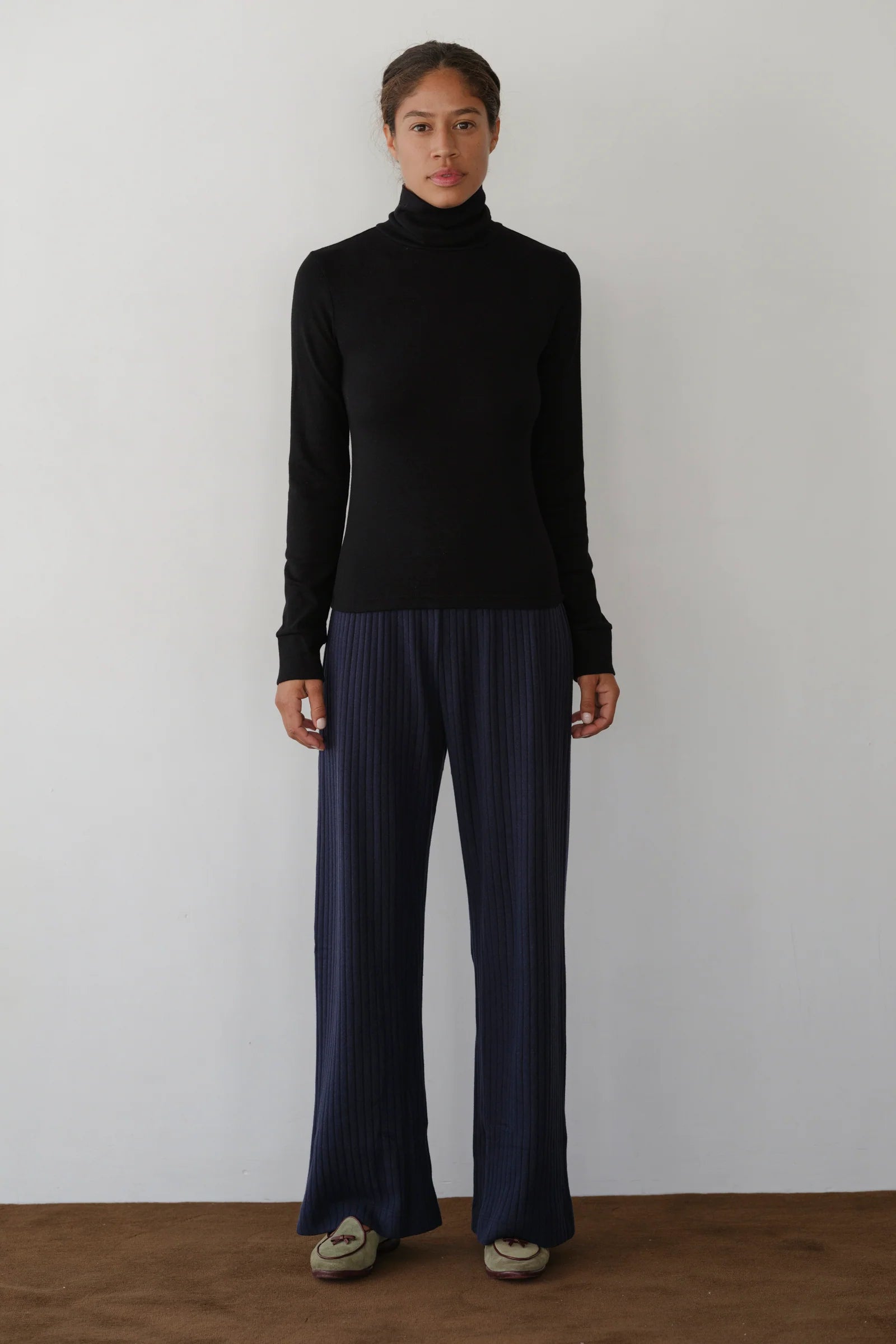 A person stands against a plain white wall wearing Donni's The Baby Rib Turtleneck, loose-fitting dark blue ribbed pants, and beige shoes, facing forward with a neutral expression.
