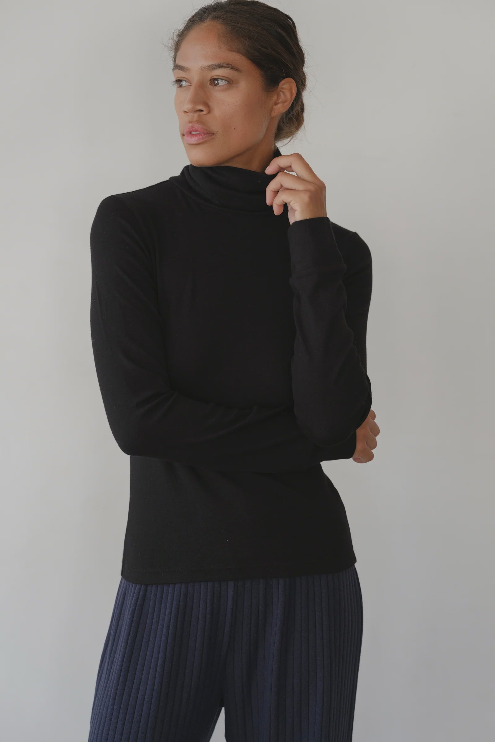 Wearing Donni's The Baby Rib Turtleneck in black and navy pleated pants, a woman stands against a plain light background, gazing to the side with one hand thoughtfully touching her chin.