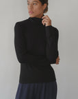 Wearing Donni's The Baby Rib Turtleneck in black and navy pleated pants, a woman stands against a plain light background, gazing to the side with one hand thoughtfully touching her chin.