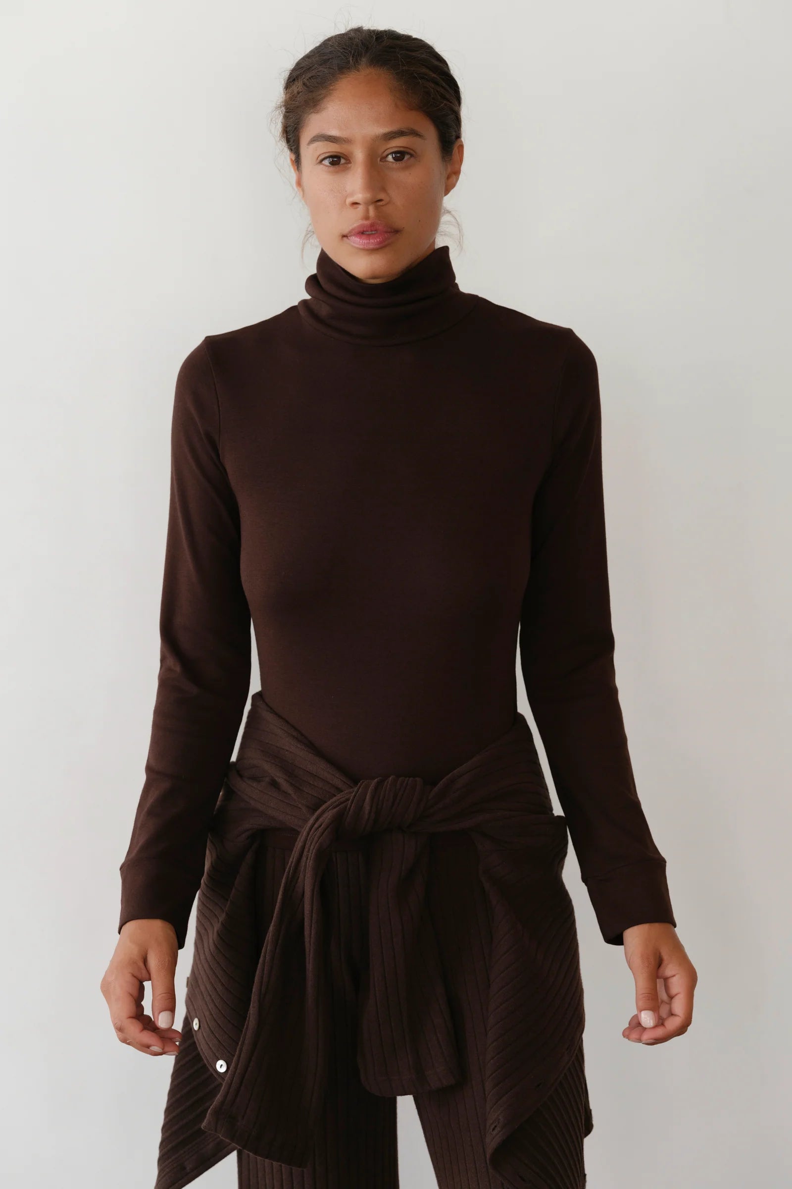 A woman stands against a plain light background, wearing Donni's The Baby Rib Turtleneck with a matching sweater tied at her waist and brown pants. She looks straight ahead with a neutral expression.