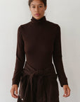 A woman stands against a plain light background, wearing Donni's The Baby Rib Turtleneck with a matching sweater tied at her waist and brown pants. She looks straight ahead with a neutral expression.