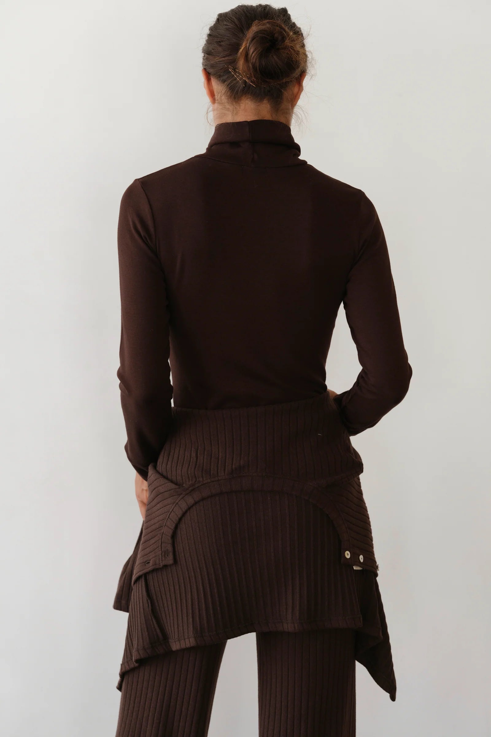 A woman with her hair in a loose bun is seen from the back, wearing Donni’s The Baby Rib Turtleneck in dark brown, paired with a ribbed knit skirt featuring layered details and matching brown ribbed pants.