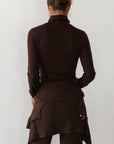 A woman with her hair in a loose bun is seen from the back, wearing Donni’s The Baby Rib Turtleneck in dark brown, paired with a ribbed knit skirt featuring layered details and matching brown ribbed pants.