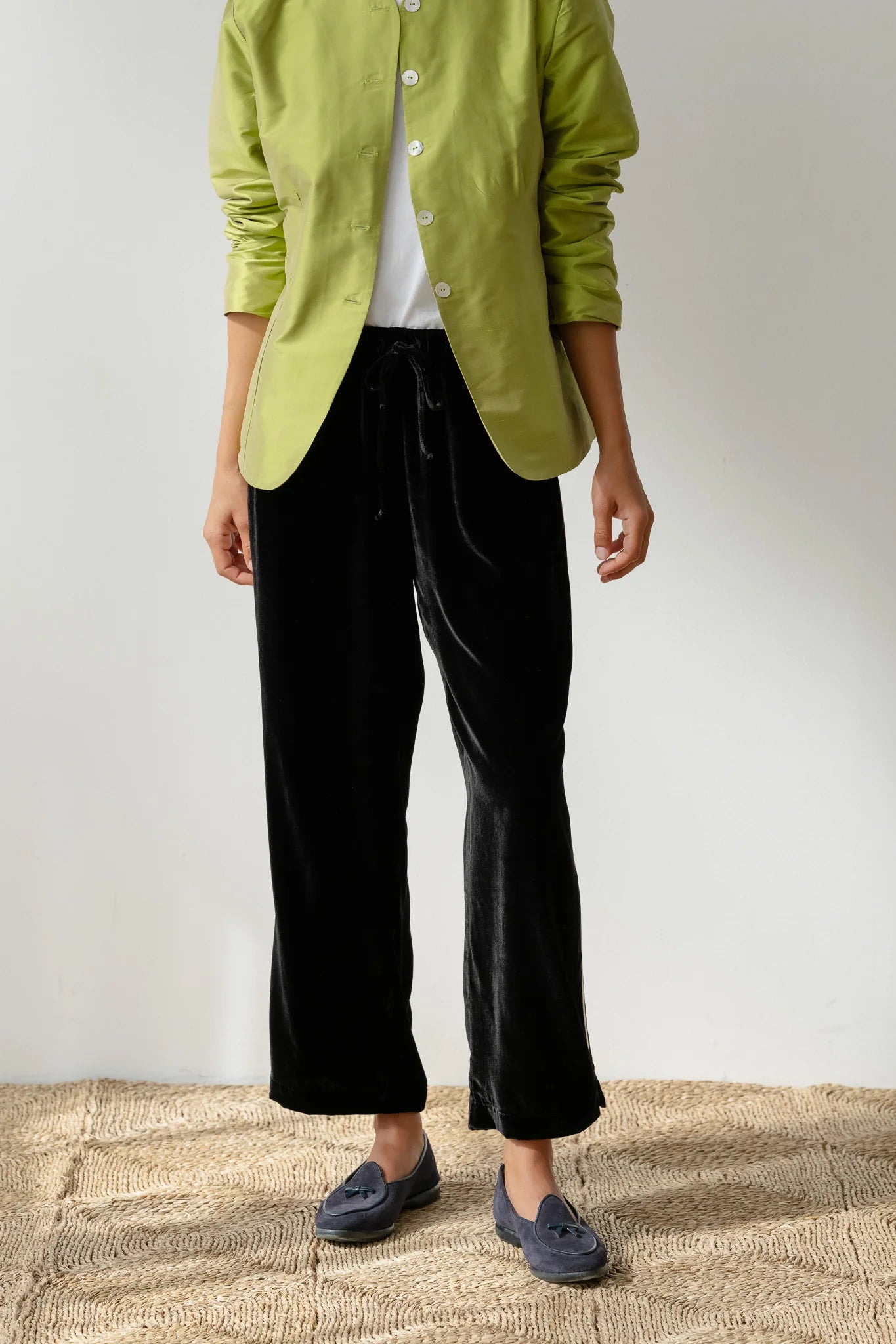 Wearing a light green jacket over a white top, someone pairs Donni's The Velvet Stripe Crop pants in a relaxed fit with a drawstring waistband and gray loafers. A textured beige background complements the outfit while keeping their head out of view.