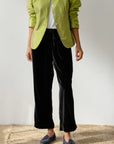 Wearing a light green jacket over a white top, someone pairs Donni's The Velvet Stripe Crop pants in a relaxed fit with a drawstring waistband and gray loafers. A textured beige background complements the outfit while keeping their head out of view.