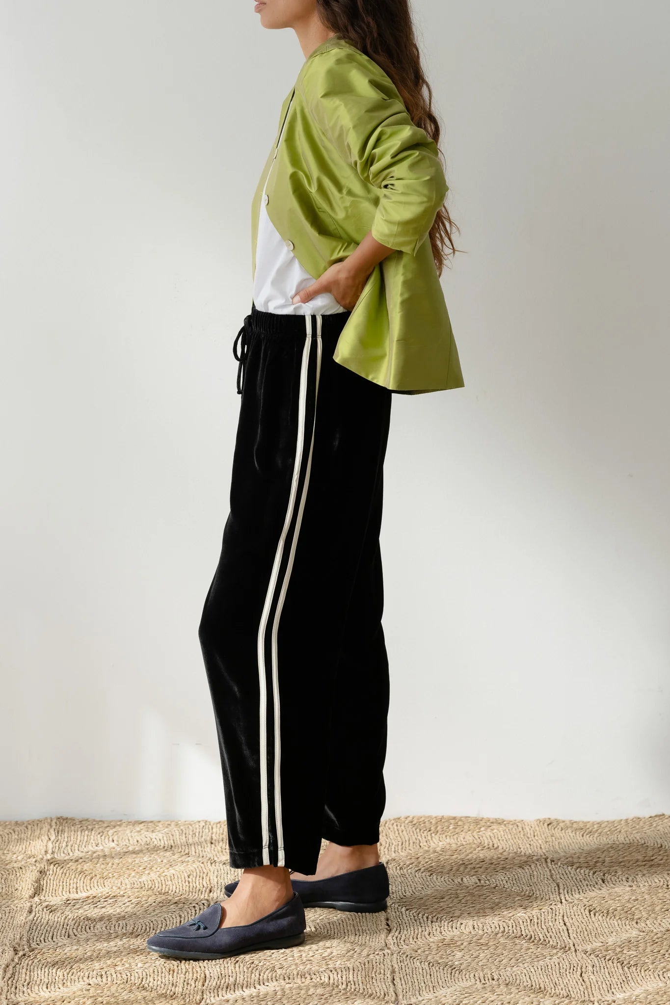 In a profile pose against a plain backdrop, someone wears The Velvet Stripe Crop by Donni with a green jacket and white shirt. Relaxed fit pants are paired with dark slip-on shoes, hand in pocket, showcasing casual elegance.