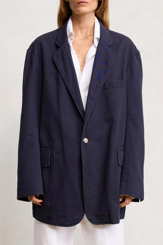 A person wears the Denimist DECONSTRUCTED BLAZER—an oversized navy blue jacket with large lapels and front pockets—over a white shirt and pants, standing against a plain light background. The face is partially out of frame.