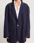 A person wears the Denimist DECONSTRUCTED BLAZER—an oversized navy blue jacket with large lapels and front pockets—over a white shirt and pants, standing against a plain light background. The face is partially out of frame.