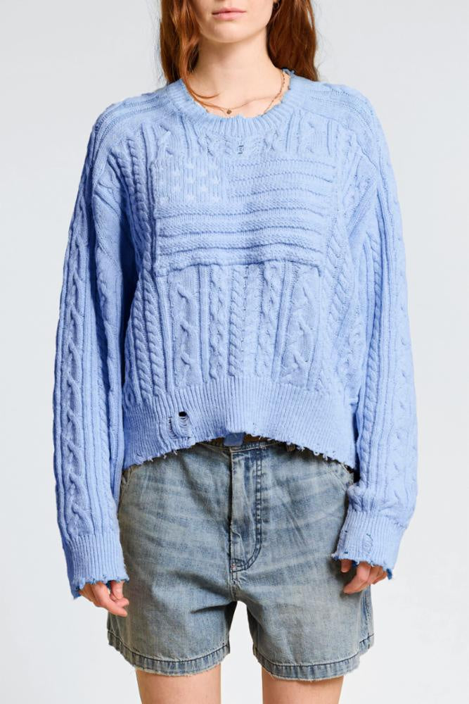 Wearing the Denimist O/S Crop Flag Sweater, an oversized blue cotton piece with frayed edges, and light denim shorts, the model stands against a plain background.