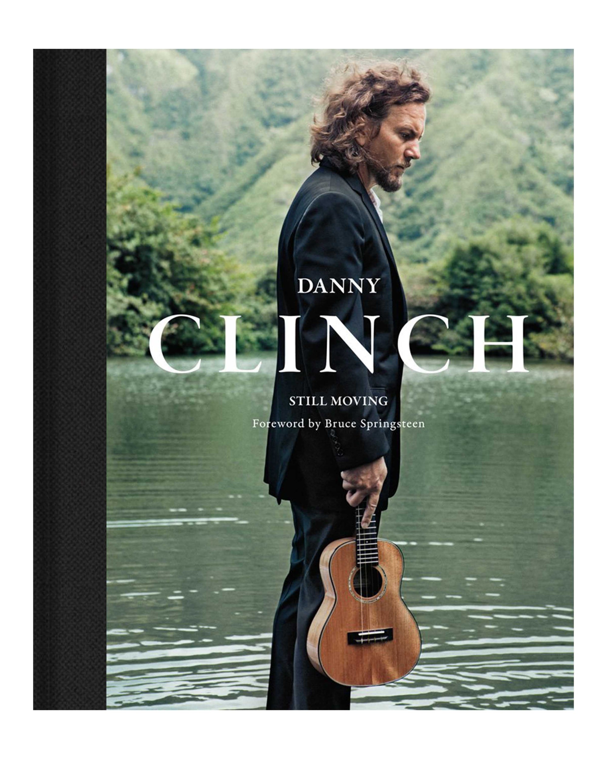 A man in a suit with a ukulele stands by a calm lake before green mountains, reflecting the spirit of music photography in "Danny Clinch" by Hachette Book Group. Foreword by Bruce Springsteen.