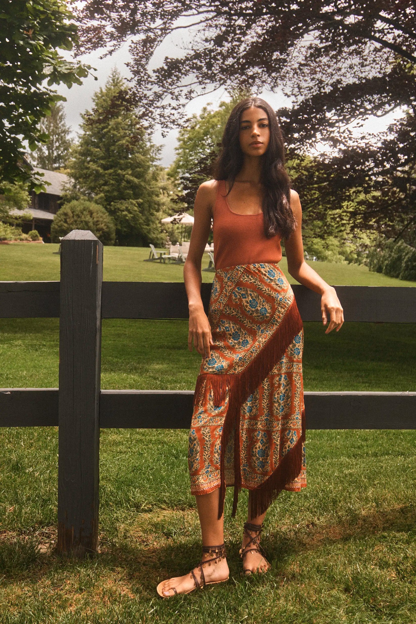 Hunter Bell NYC Allyne Skirt with fringe in rust, styled outdoors with a tank top and sandals by a wooden fence.