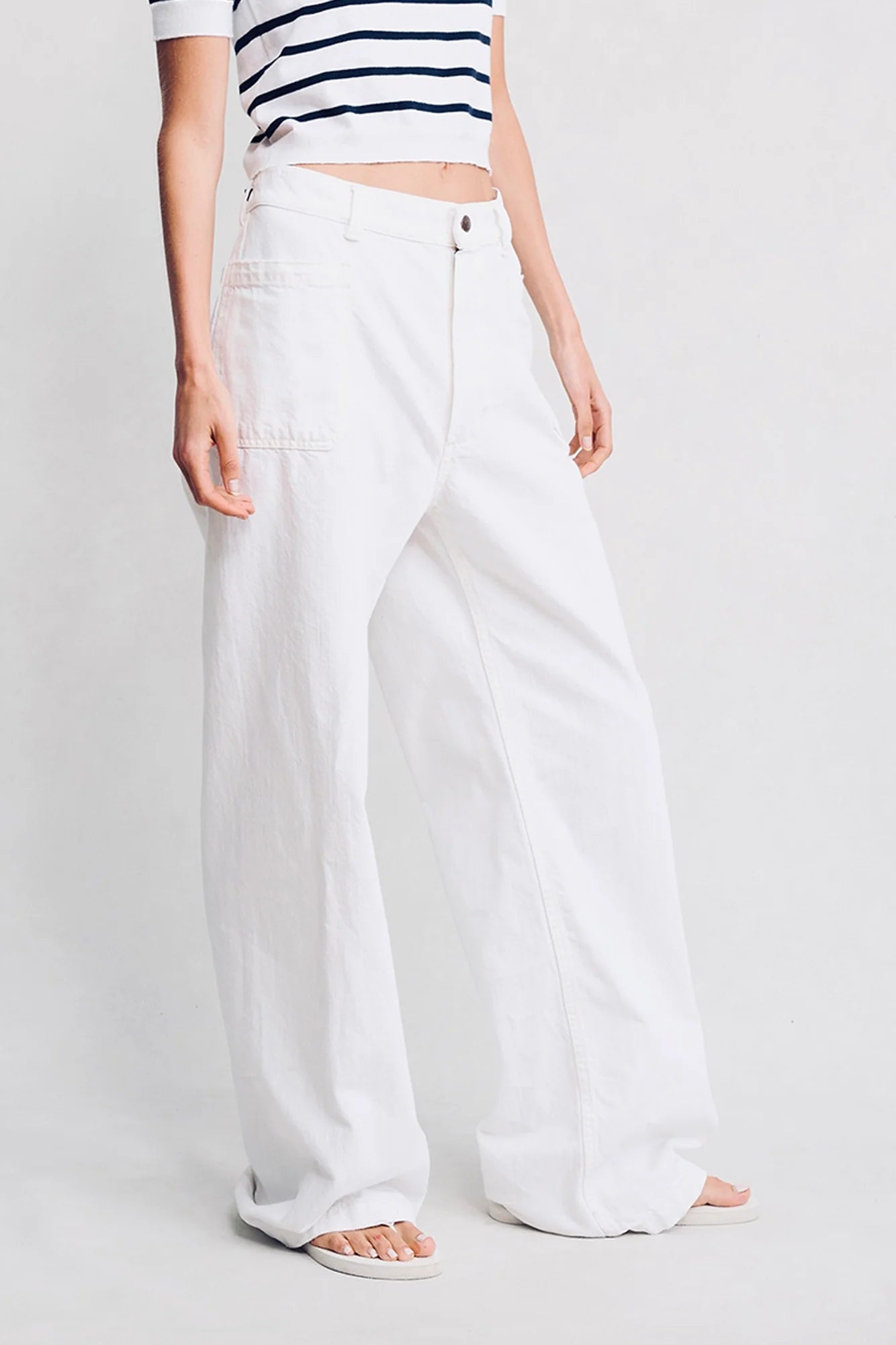 A person models Denimist’s DECK PANT WHITE WASH in white denim, paired with a striped short-sleeve top and white sandals, standing against a plain light background. The image is cropped below the shoulders.