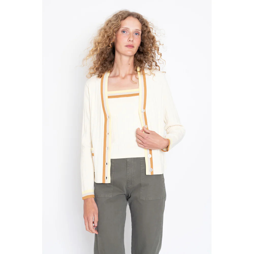 A person with curly hair models Kule's The Dede Cream/Hazelnut cardigan, featuring yellow trim, over a matching top and olive green pants. They face the camera against a white background, perfectly capturing a spring/summer vibe.